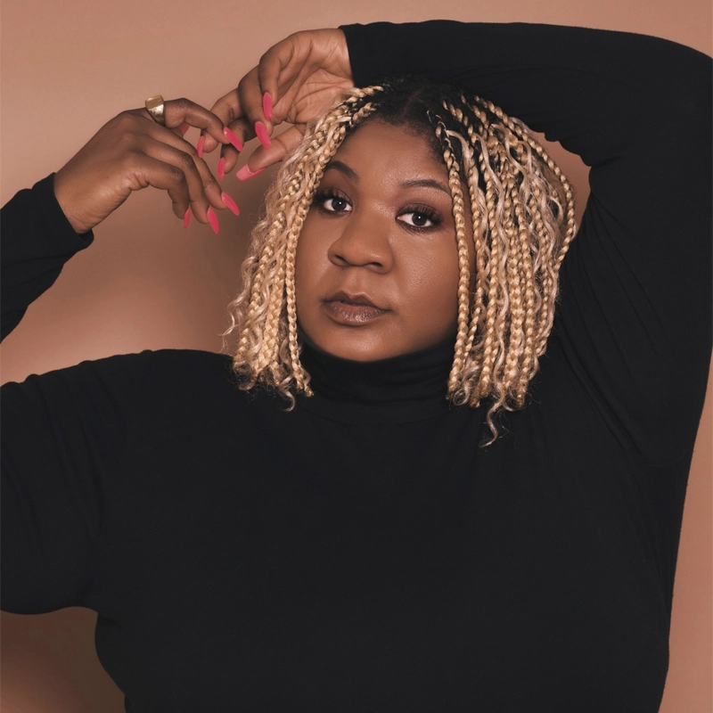 Jazmin McCray, with blond box braids, poses confidently against a brown background. She wears a black turtleneck and shows off long pink nails—one hand above her head, the other touching her braids. Her expression is calm and self-assured.