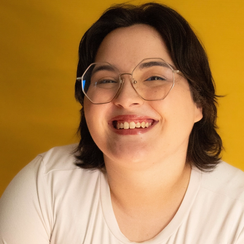 Gabby Medeck, with short dark hair and glasses, smiles warmly at the camera. Wearing a light-colored top against a solid yellow background, the soft lighting gives this portrait a welcoming, cheerful tone.