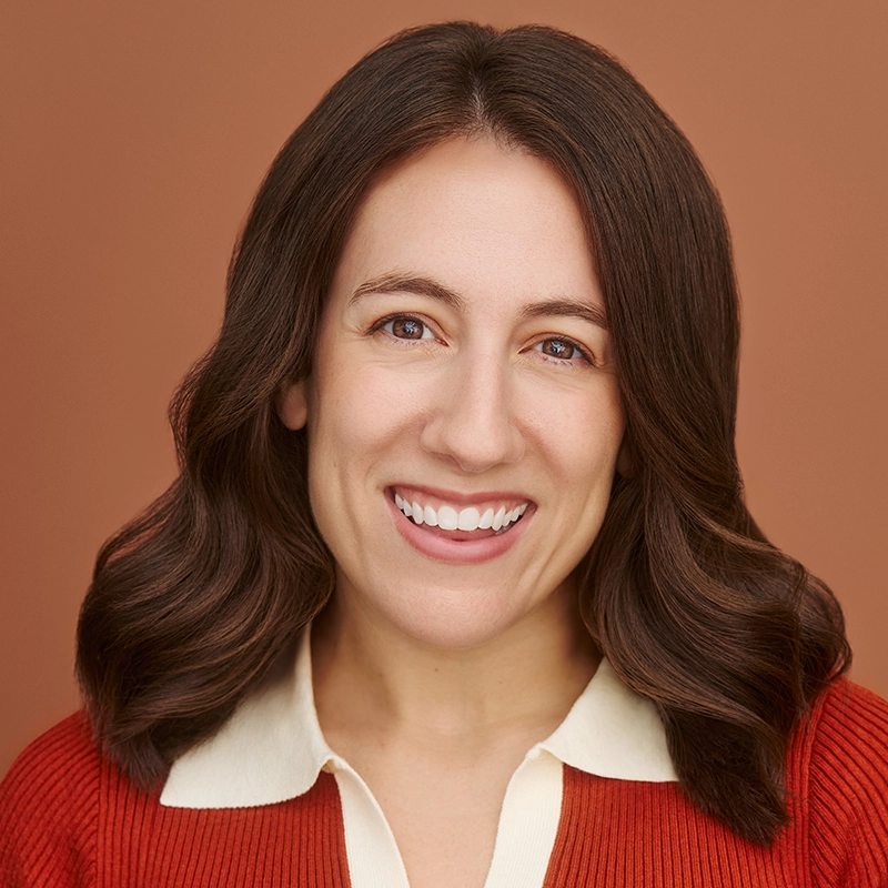 Lyla Karekinian, with long, wavy brown hair, smiles warmly at the camera. She wears a cream collared shirt under a rust-colored sweater, set against a soft, solid light brown background for a professional portrait.