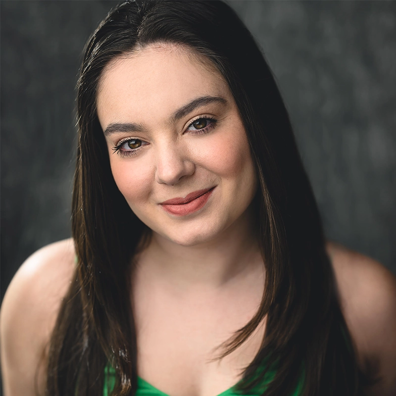 Melissa Aliotta, a young woman with long, straight dark brown hair, smiles gently. She wears a sleeveless green top. The background is dark and softly blurred, drawing focus to her warm expression and natural makeup.