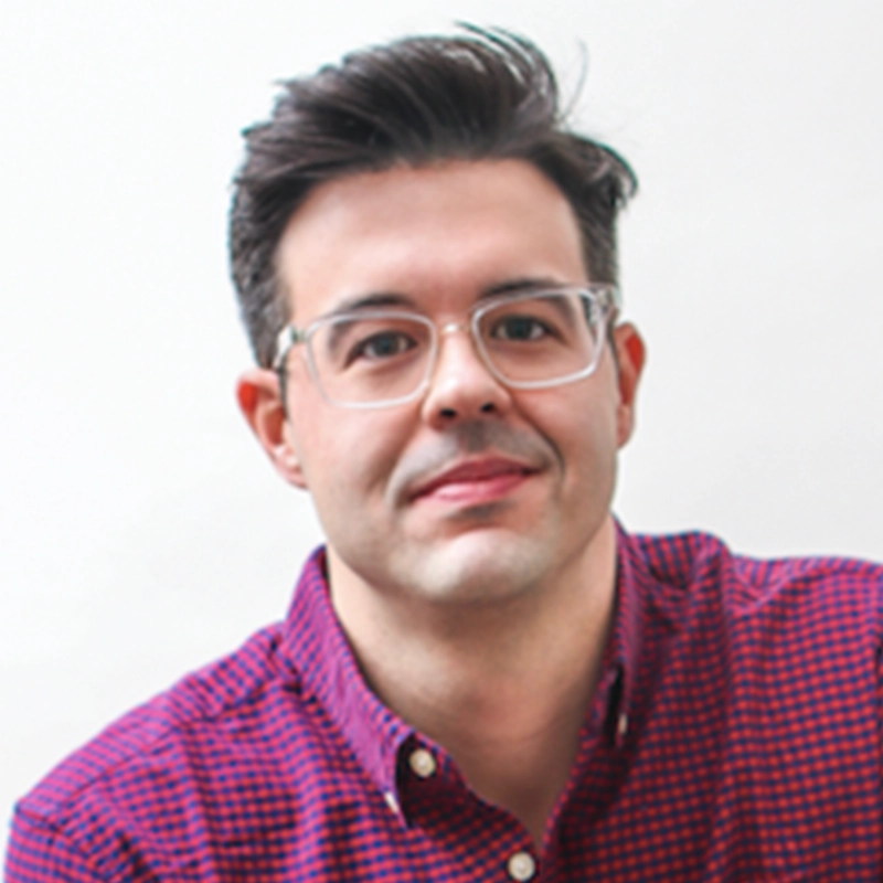 Michael Bello, a man with short dark hair, clear-framed glasses, and a red and blue checkered shirt, is smiling softly in front of a plain, light-colored background.