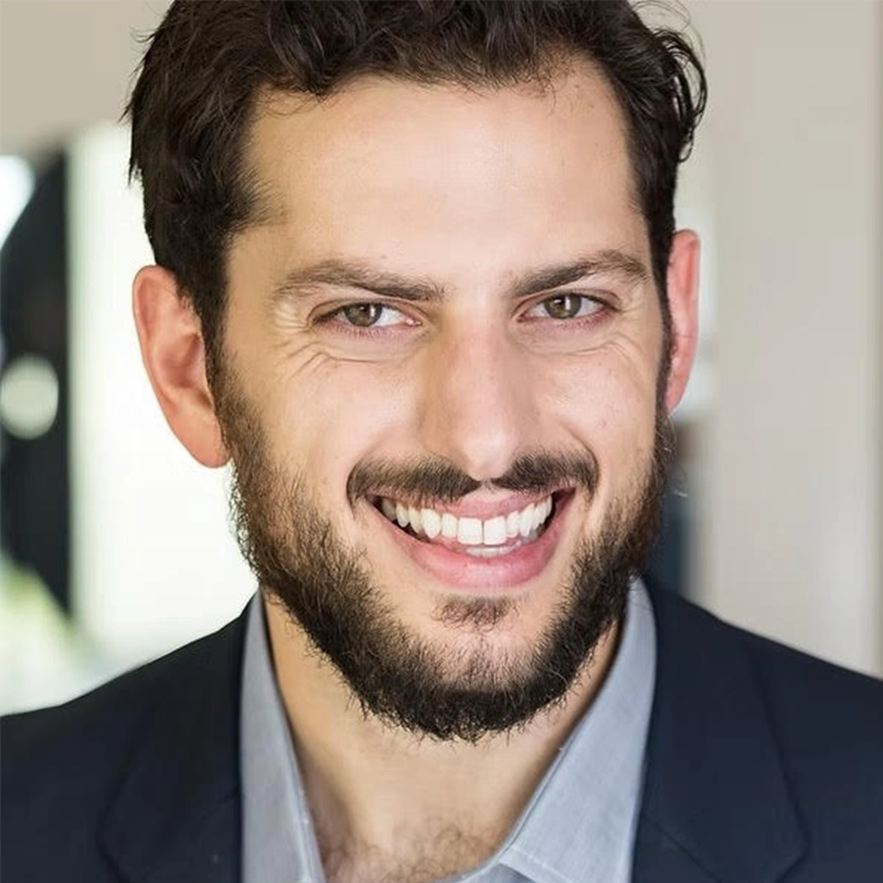 A man with short, dark hair and a full beard smiles warmly at the camera. He is wearing a navy blazer over a light gray collared shirt. The softly blurred background features hints of indoor lighting and neutral tones, photographed by Abby Wolanin.