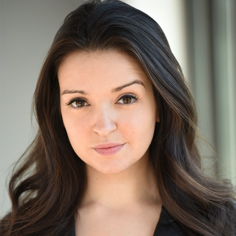 Aidan Michael Kelly captures a young woman with long, dark brown hair and fair skin facing the camera. She has brown eyes, subtle makeup, a neutral expression, and wears a black top against a softly blurred background with neutral tones.