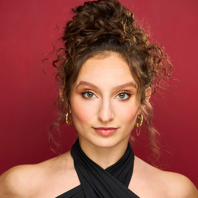A young woman with curly brown hair in a loose updo poses against a deep red background. She wears gold hoop earrings, natural makeup with winged eyeliner, and a black halter top. Her expression is calm and confident, facing directly toward the camera.