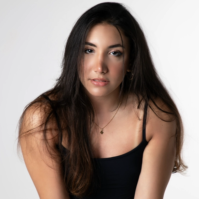 Melissa Milano, a young woman with long dark hair and light skin, wears a black tank top and delicate necklace, gazing softly at the camera against a plain white background. Her makeup is subtle, with natural tones and a slight sheen on her lips.