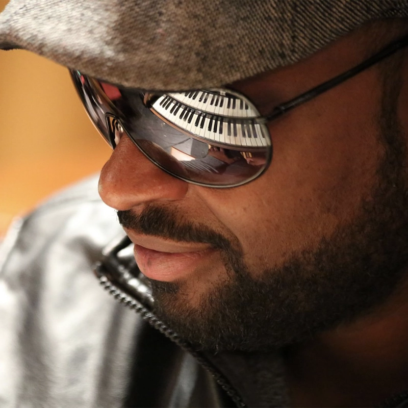 A close-up of Michael O. Mitchell wearing a gray cap and reflective sunglasses. The reflection in his sunglasses shows a piano keyboard. He has a neatly trimmed beard, mustache, and is dressed in a dark leather jacket, looking relaxed and focused.