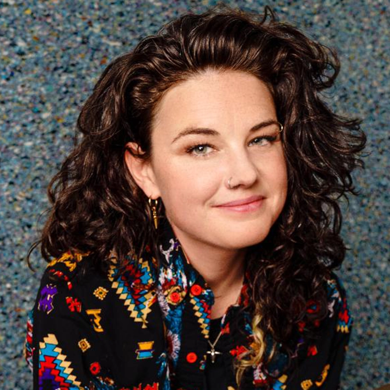 Francesca Pastore, a woman with curly brown hair, a nose ring, and hoop earrings, smiles softly. She wears a black shirt with bright geometric patterns. Against a blue, speckled background, her head is slightly tilted as she looks at the camera.