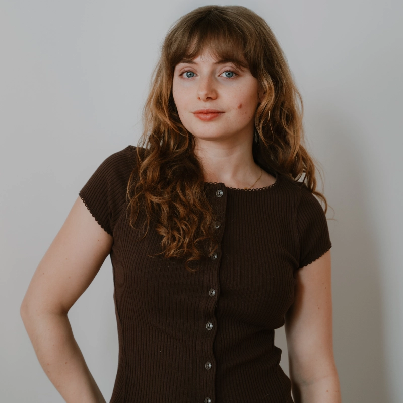 A young woman with long, wavy light brown hair and bangs stands against a plain light-colored wall. She wears a short-sleeved, ribbed, dark brown top with buttons down the front and looks calmly at the camera with a neutral expression.