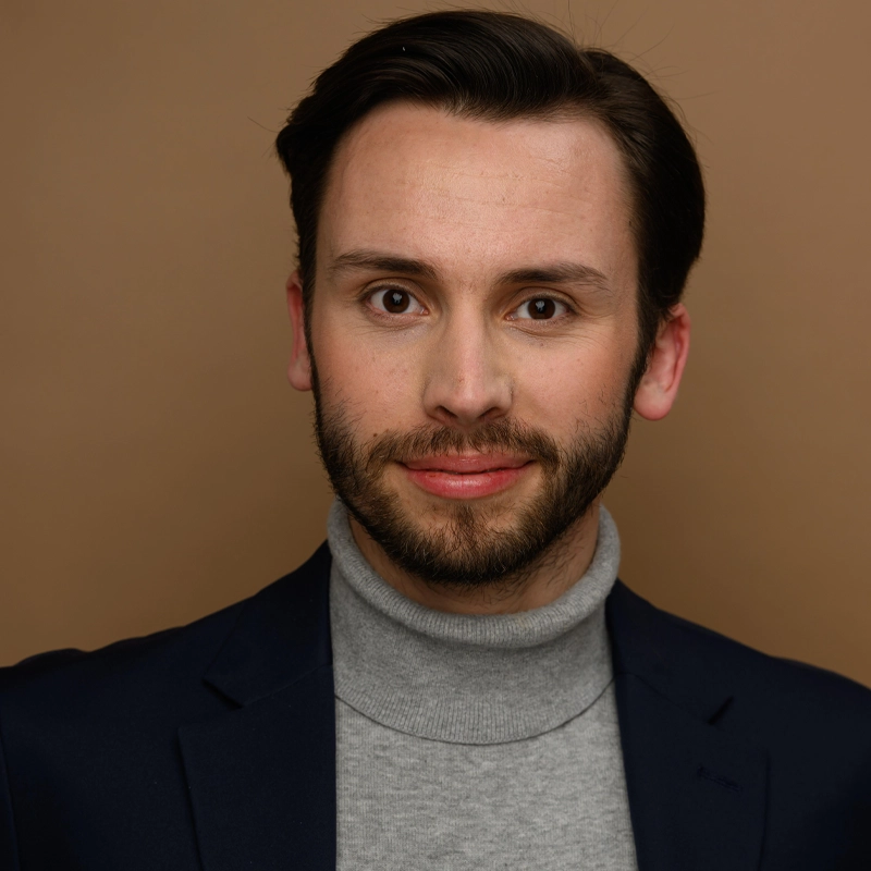 Jack F. Murphy, a man with light skin, short dark hair, and a trimmed beard, smiles slightly. He wears a navy blazer over a light gray turtleneck sweater against a plain warm brown background. Even lighting highlights his face.