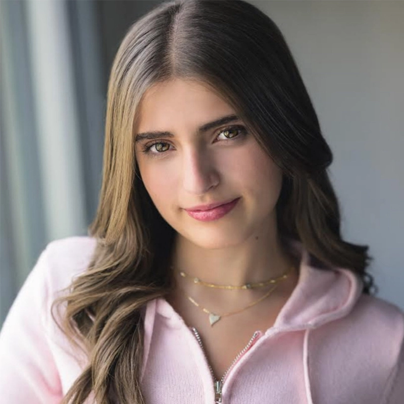 Lyla Karekinian, a young woman with long brown hair, wears a light pink zip-up hoodie and layered gold necklaces as she stands by a window. She gazes at the camera with a slight smile, her face softly highlighted against the blurred background.