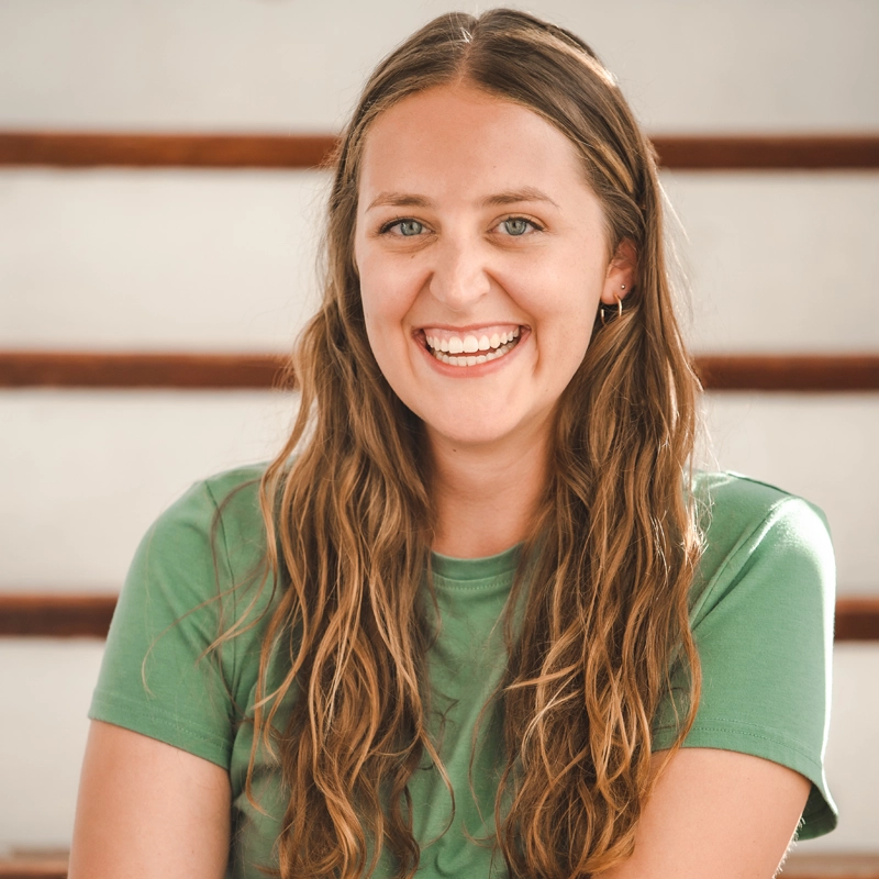 Sara Naumann, a young woman with long, wavy brown hair and blue eyes, smiles warmly. She wears a light green t-shirt and small hoop earrings. The softly blurred background features wooden horizontal lines, possibly stairs or rails.