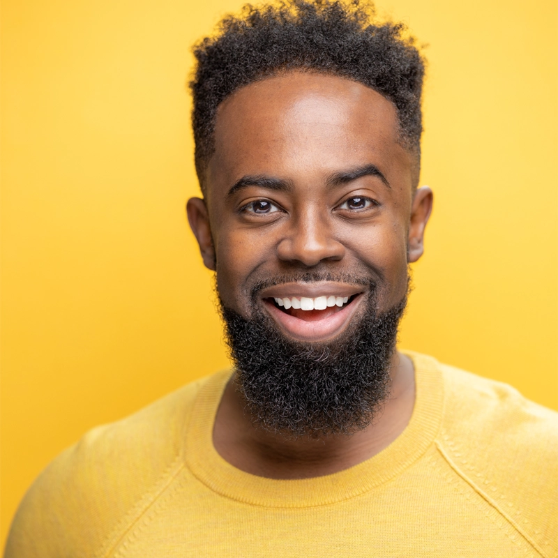 A man with dark skin, a short curly haircut, and a full beard smiles broadly. Justin G. Nelson wears a yellow sweater, and the solid yellow background creates a bright and cheerful atmosphere.