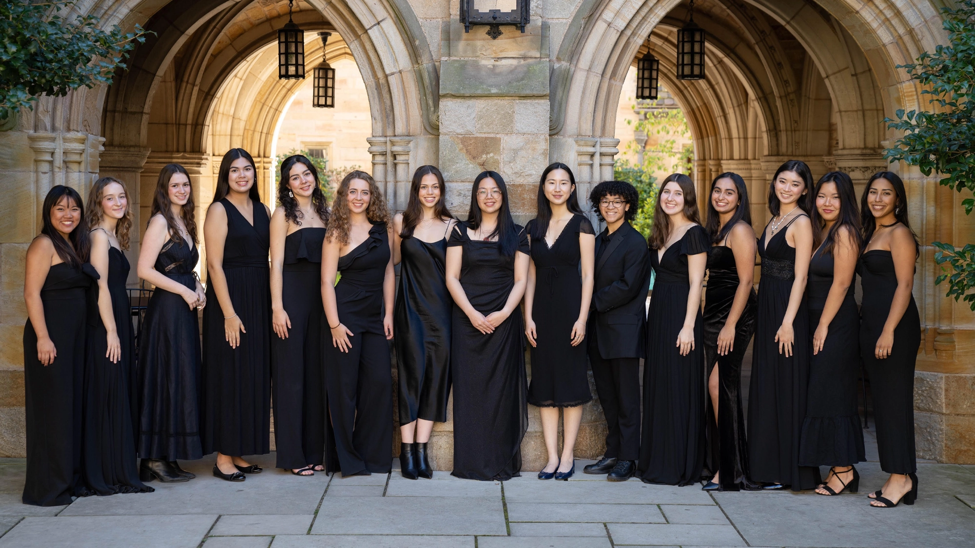 Fifteen women, part of an SSAA A Capella group, stand side by side in front of stone archways at Yale University, all in formal black dresses except one in a suit. They smile at the camera, framed by ivy and lanterns on a stone courtyard.