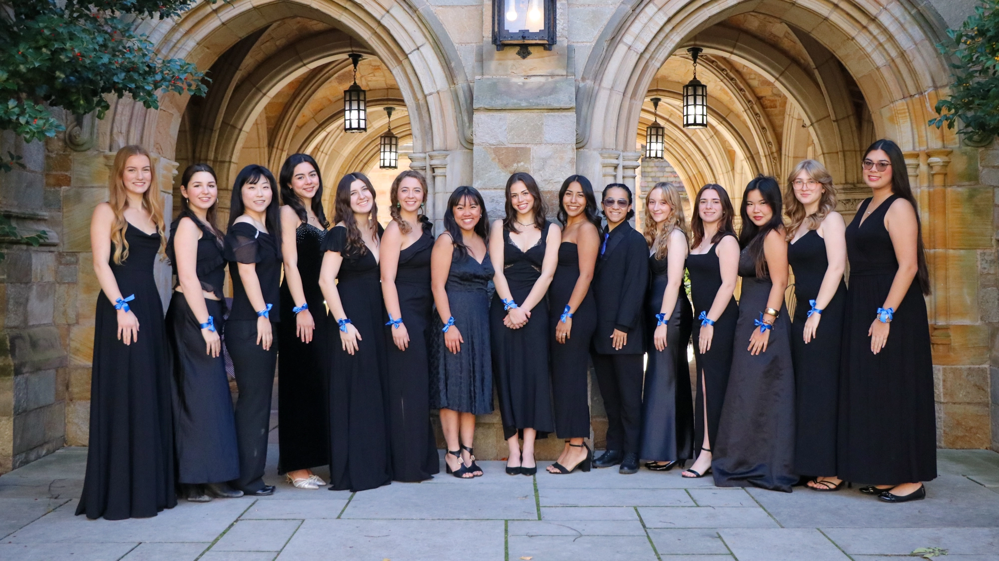 New Blue of Yale- Show Image Fourteen young women in long black dresses, members of an SSAA A Capella group, stand smiling with arms linked before a stone archway lit by lanterns. All wear blue wristbands, suggesting a formal Yale University event in an elegant historic setting.