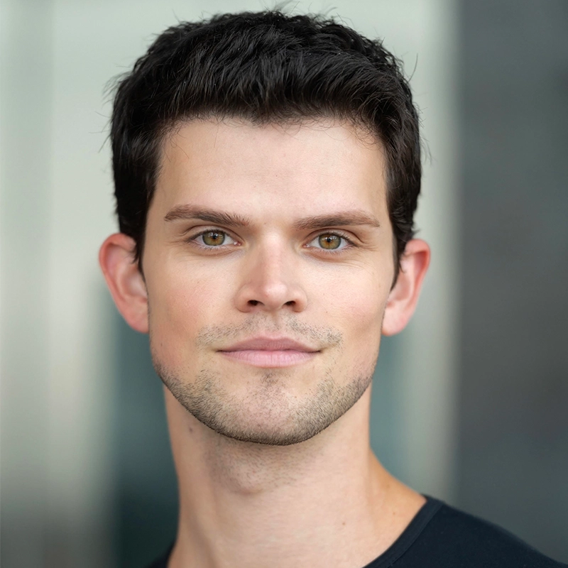 A young man with short, dark hair and fair skin looks directly at the camera with a slight smile. He wears a black shirt. The background is softly blurred in grey tones, putting focus on his face.
