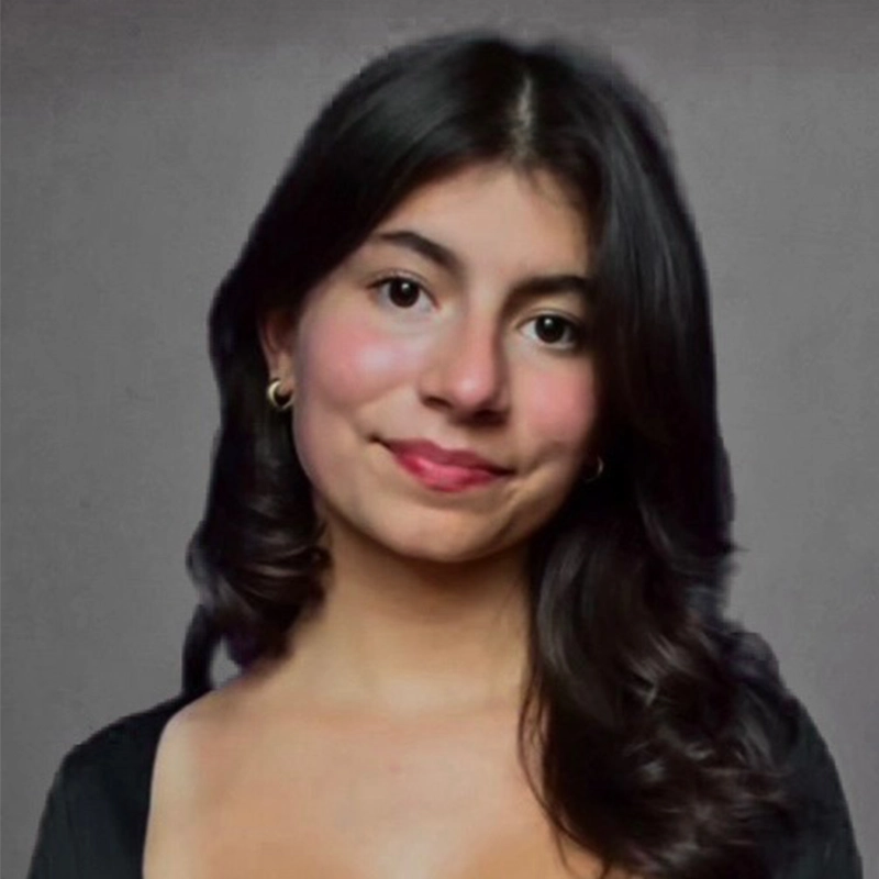 Sara-Louise Oakes, a young woman with long, wavy dark hair, smiles softly at the camera. She wears gold hoop earrings and a black top with a low neckline. Her background is plain gray and her skin has a warm, natural tone.
