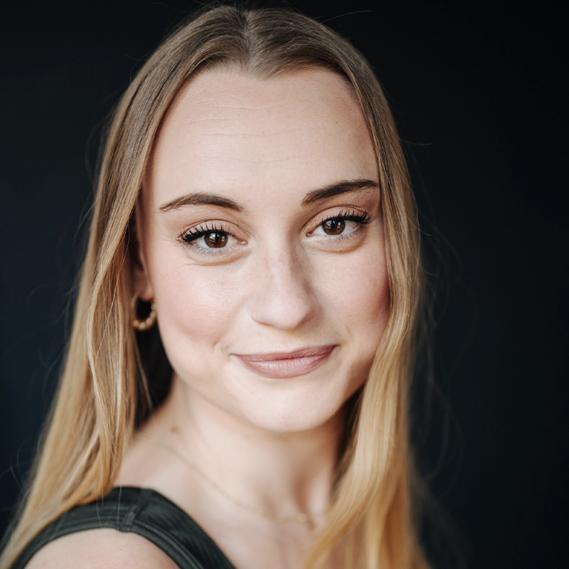 A young woman with long straight blonde hair, light skin, and brown eyes smiles softly at the camera. She wears natural makeup, a sleeveless dark green top, small hoop earrings, and a delicate necklace, set against a dark, plain background.
