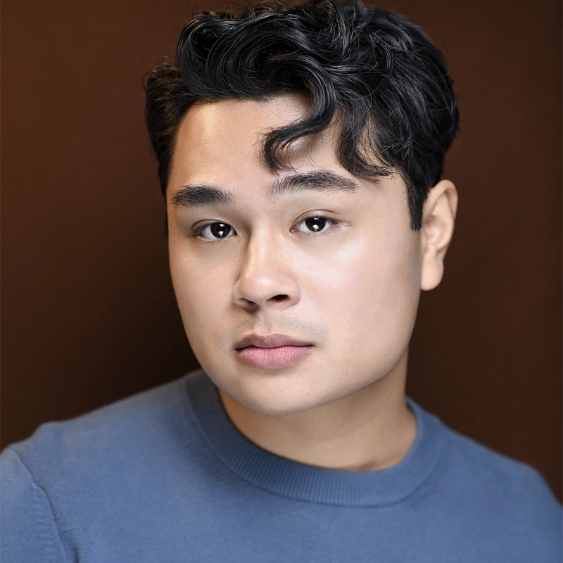 A young person with short, wavy black hair and fair skin wears a blue crew-neck shirt. Gabriel Vernon Nunag looks directly at the camera with a neutral expression, set against a solid dark brown background that highlights their features.