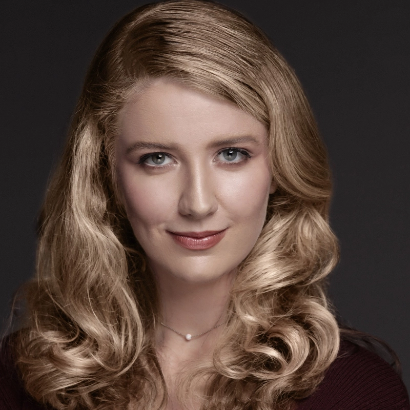 Grace O'Keefe, with long, wavy blonde hair, poses against a dark background. She has fair skin, blue eyes, and a slight smile. She wears a dark V-neck top and a delicate necklace with a small round pendant.