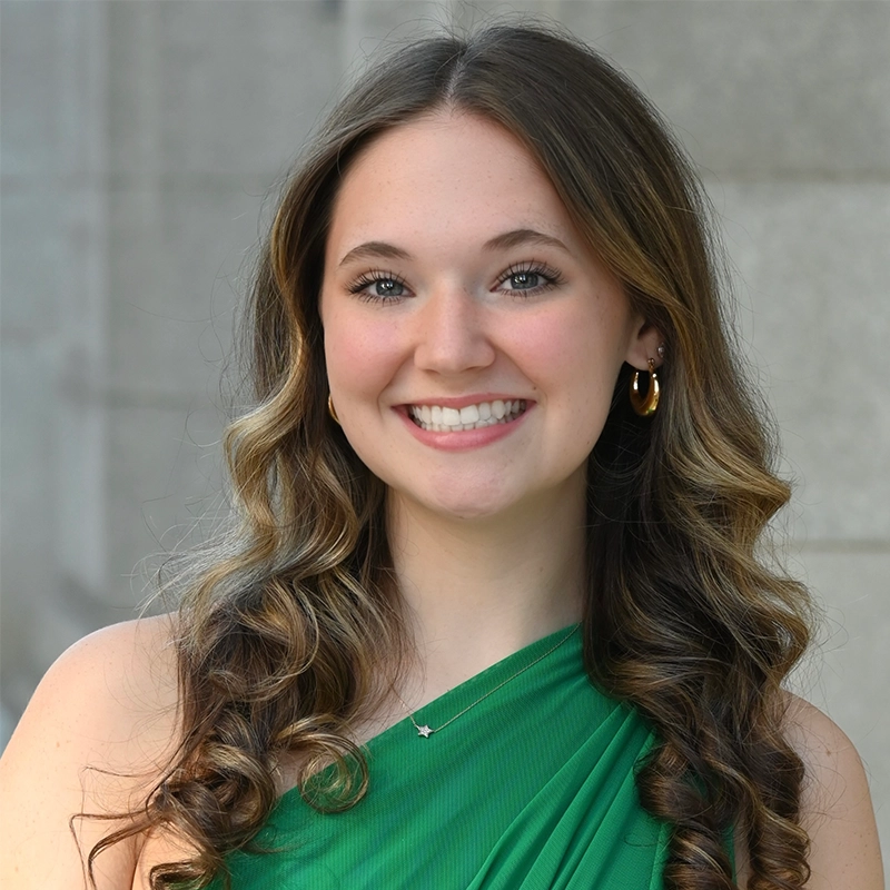 Sara-Louise Oakes, a young woman with long, wavy brown hair and light highlights, smiles at the camera. She wears a sleeveless green dress, gold hoop earrings, and a delicate necklace against a blurred stone wall, adding a professional touch.