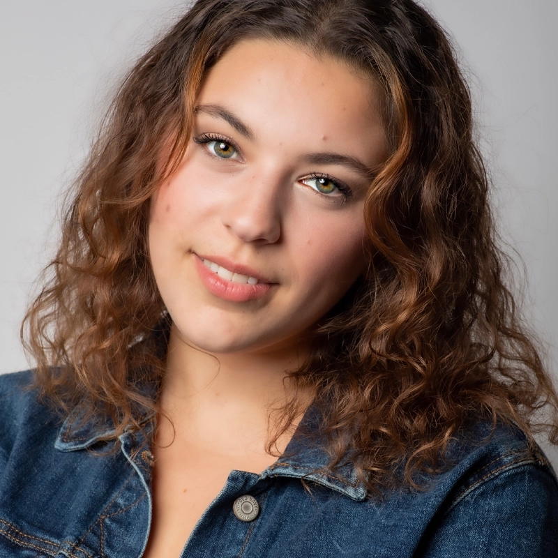 Taylor Parker, a young woman with wavy brown hair, smiles softly at the camera. She is wearing a denim jacket over a light top. The plain, light gray background keeps the focus on her face and natural expression.