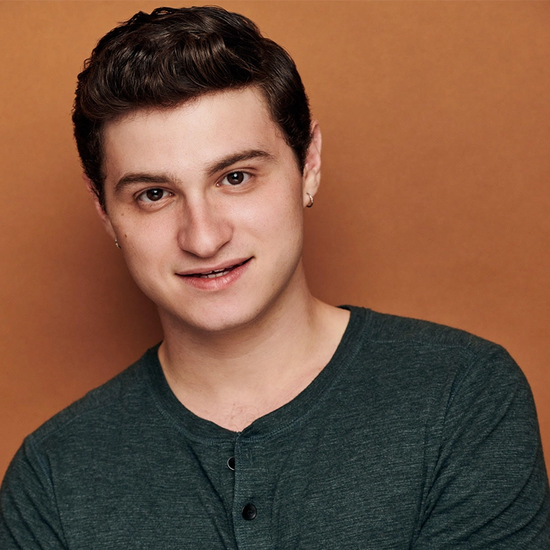 A young person with short, wavy brown hair and fair skin smiles gently at the camera. They wear a dark green henley shirt and a small hoop earring in one ear. The background is a solid warm brown color.