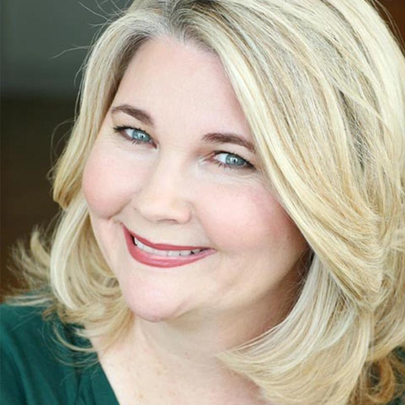 A smiling middle-aged woman with fair skin and shoulder-length blonde hair. She is wearing a green top and has light blue eyes. The background is softly blurred, drawing focus to her friendly expression and subtle makeup.