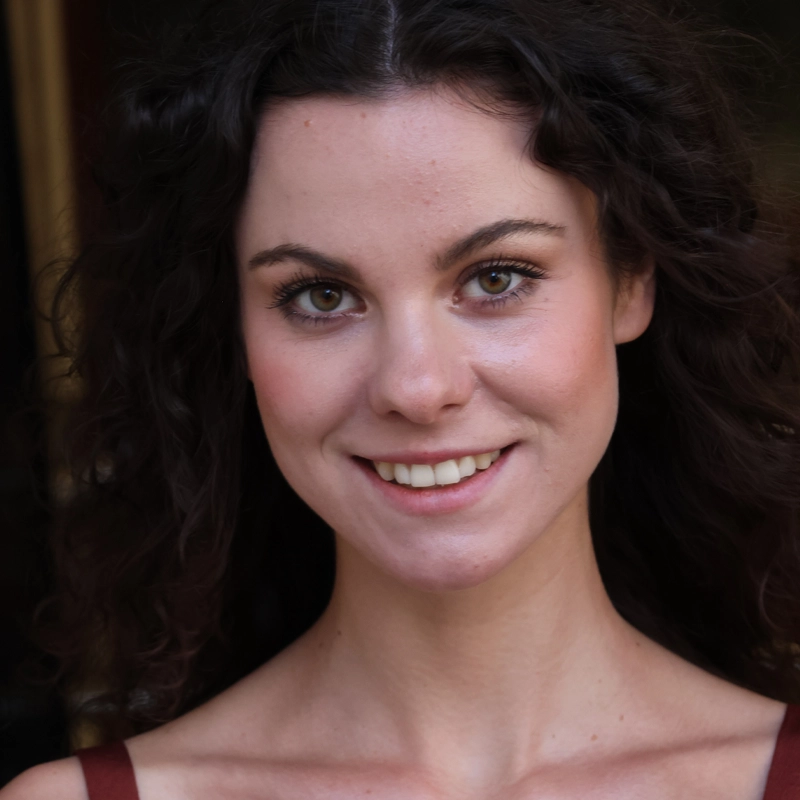 A young woman with fair skin and curly dark brown hair smiles at the camera. She has brown eyes, natural makeup, and is wearing a sleeveless brown top. The background is dark and out of focus, drawing attention to her face.