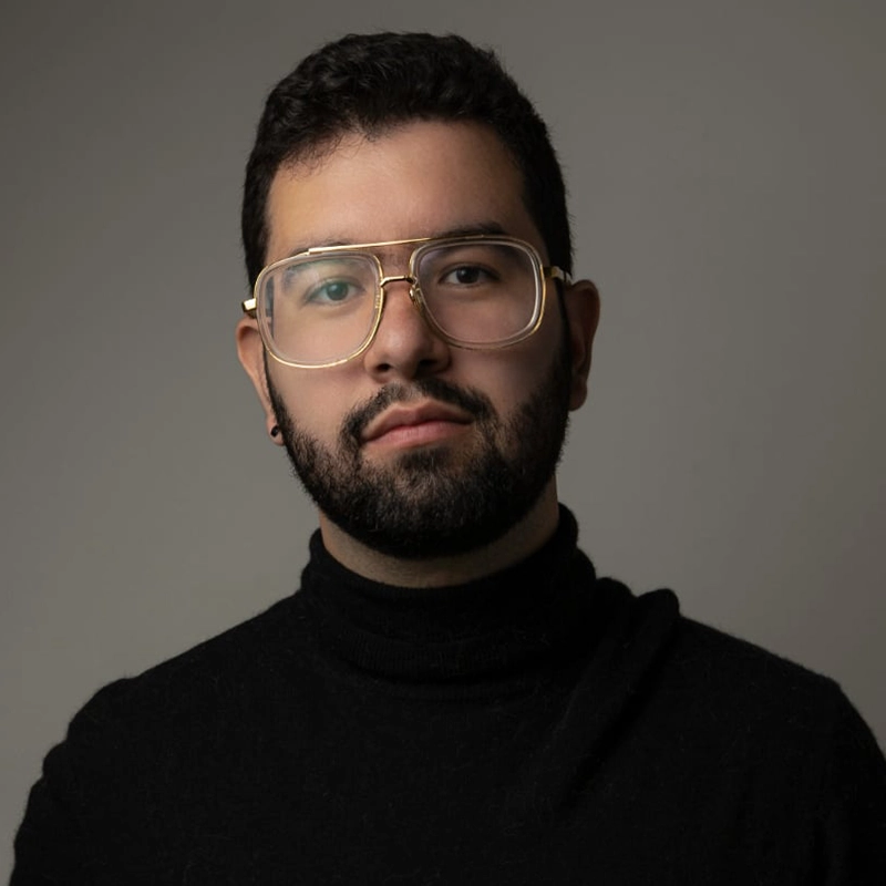 A man with short, dark hair and a trimmed beard faces the camera. He wears large, gold-rimmed eyeglasses and a black turtleneck sweater. The background is plain and neutral, with soft lighting highlighting his face.