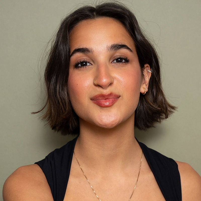 A young woman named Kelly with short, dark brown hair smiles softly at the camera. She wears a black top, a gold nose ring, small hoop earrings, and a delicate chain necklace. Her light skin is complemented by groomed brows and rosy cheeks against a beige background.