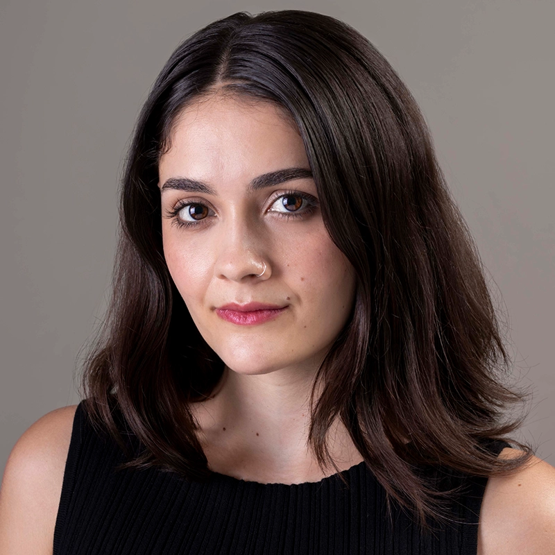 A young woman, Abby Wolanin, with medium-length dark brown hair parted slightly to the side, wearing a sleeveless black top. She has a small nose ring and neutral makeup, looking directly at the camera with a subtle, confident expression against a plain gray background.