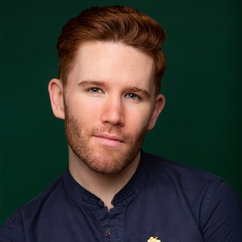 A young man named Tyler with short red hair and a trimmed beard poses against a dark green background in Raleigh. He wears a navy blue button-up shirt and looks directly at the camera with a slight smile and confident expression.