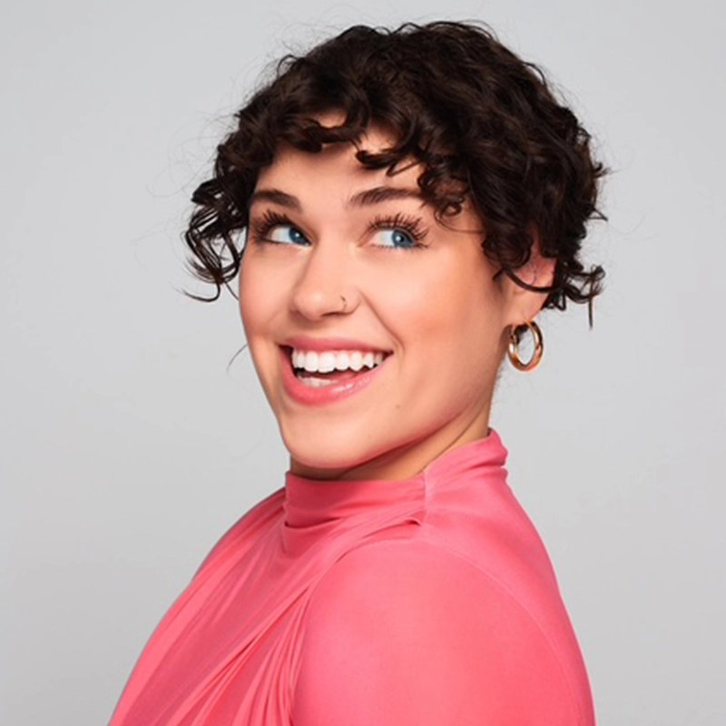 A young person with short, curly brown hair smiles brightly while looking to the side. They wear a sheer pink top and gold hoop earrings. The cheerful, vibrant mood echoes Kourtnie Dwornikowski’s signature uplifting style.