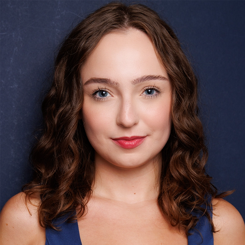 Carissa Ferguson, a young woman with fair skin, curly brown hair, blue eyes, and pink lipstick, looks directly at the camera. She wears a blue sleeveless top and poses against a dark blue background with a slight smile.