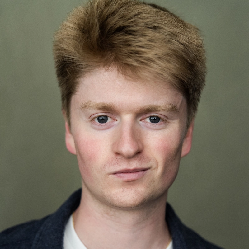 A young man with fair skin, short, thick, light brown hair, and blue eyes looks directly at the camera with a slight smile. He is wearing a white shirt under a dark jacket, and the background is plain and neutral-colored.