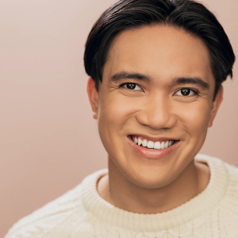 A young man, Hayden Rivas, with straight dark hair and medium skin tone smiles warmly. He wears a cream-colored knit sweater against a soft pink background. Only his head and shoulders are visible, with bright and even lighting.