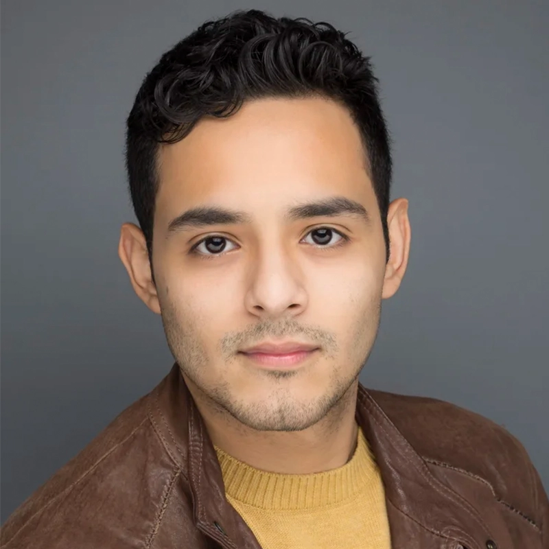A young man with short, wavy dark hair looks directly at the camera with a neutral expression. He is wearing a brown leather jacket over a mustard-yellow sweater, styled in a professional headshot reminiscent of Kenna Wilson’s signature portraits.