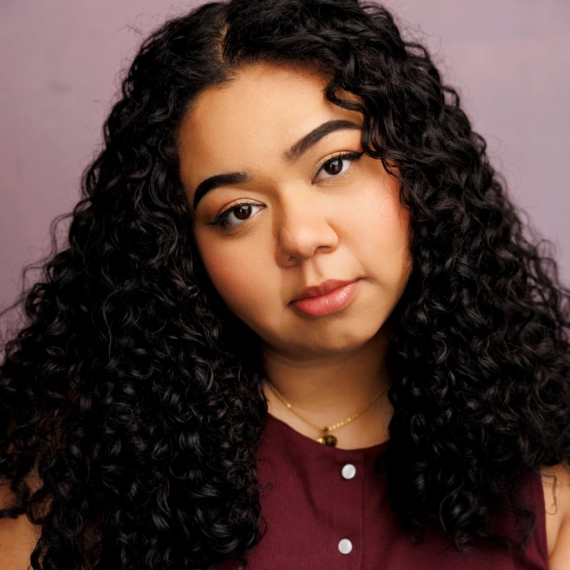 Alissa Rojas, with long, curly black hair and softly arched eyebrows, gazes calmly at the camera. She wears a burgundy sleeveless top and a gold necklace against a muted lavender background, creating a gentle and warm portrait effect.