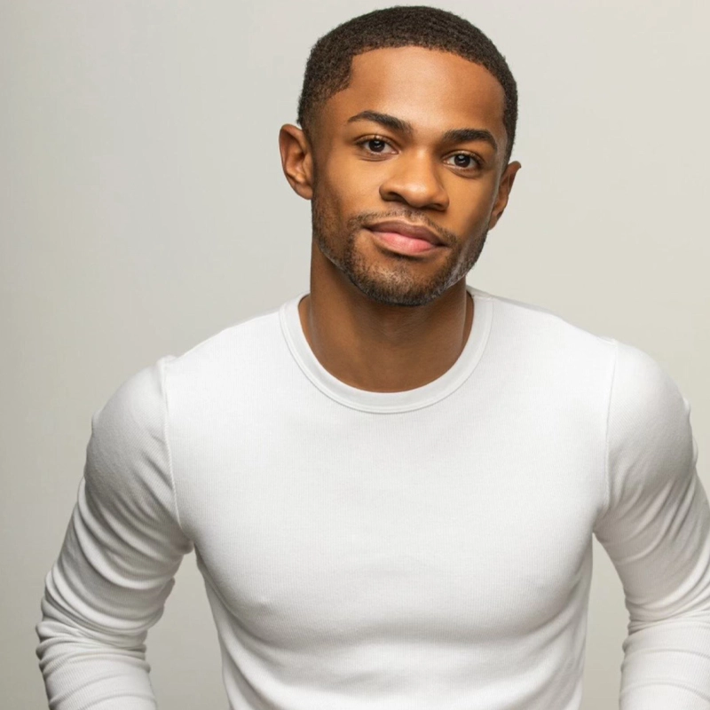 A young man with short hair and a trimmed beard smiles slightly while wearing a fitted white long-sleeve shirt. He stands against a plain, light-gray background, facing the camera with a relaxed posture and his arms by his sides.