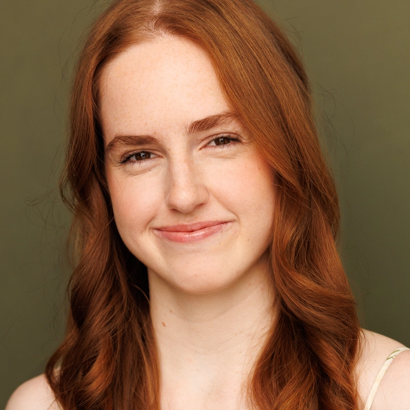 A young person with long, wavy red hair and fair skin smiles gently at the camera. They have light freckles, brown eyes, and natural makeup. The background is plain olive green, and the lighting is soft, giving a warm and friendly appearance.