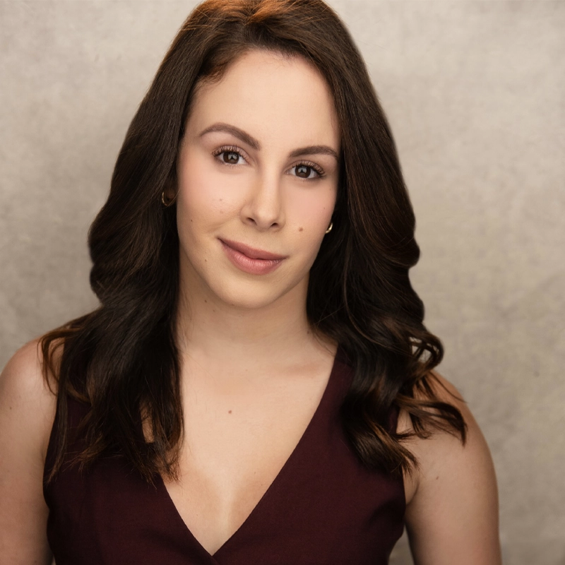 Lannie Rubio, a woman with long, wavy brown hair, smiles softly at the camera. She wears a sleeveless burgundy V-neck top and small hoop earrings. The plain, light-colored background gives the portrait a professional, studio-lit appearance.