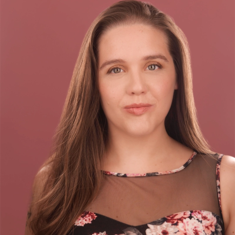 Christina Russo, a woman with long brown hair and light skin, looks at the camera with a neutral expression. She wears a sleeveless floral dress with black mesh at the top, set against a solid muted pink background.