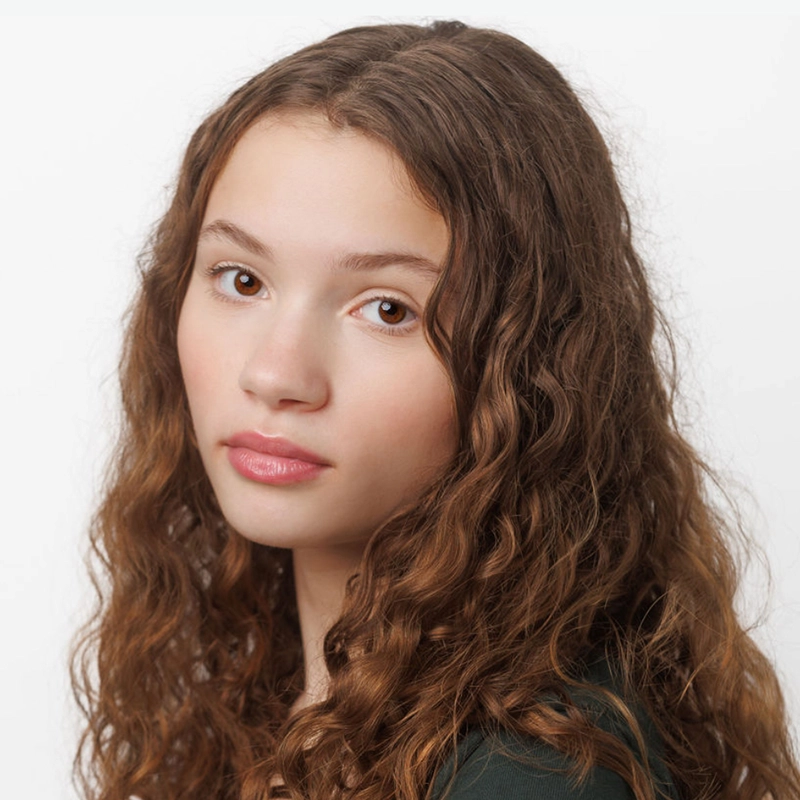 Lyla Karekinian, a young person with long, curly brown hair and fair skin, looks toward the camera with a calm expression. With light brown eyes, natural makeup, and a dark green top, their features stand out against a plain white background.