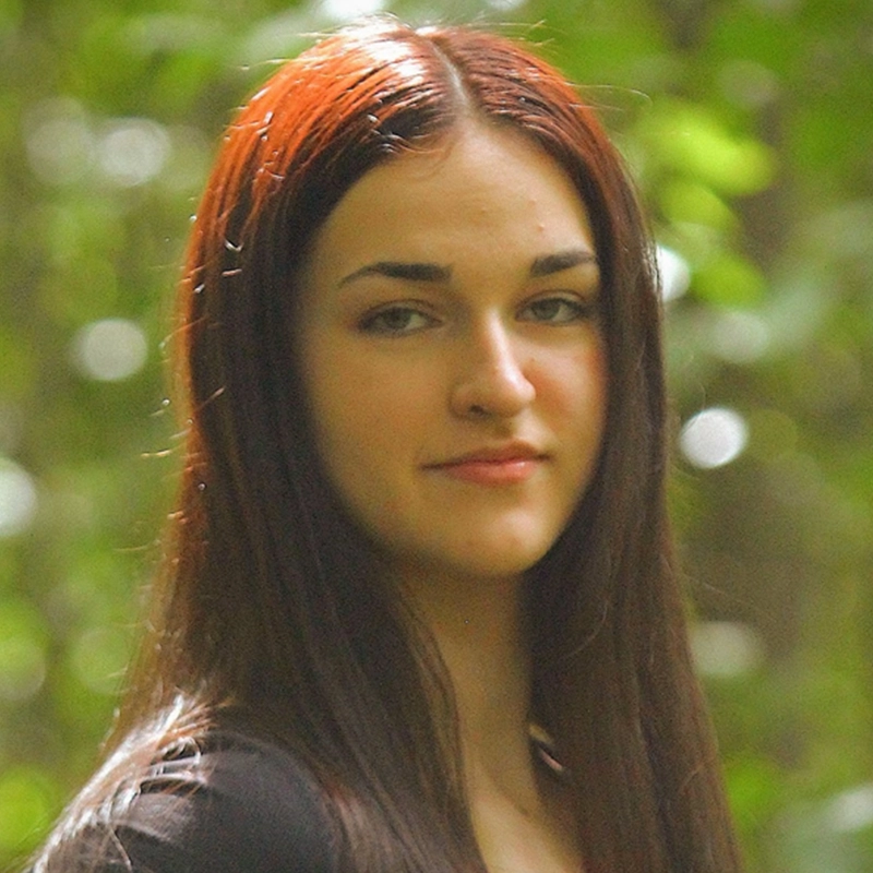 Sara-Louise Oakes, a young woman with straight, long dark brown hair, stands outdoors. She has fair skin and a slight smile, framed by blurred greenery and soft sunlight filtering through the wooded setting.