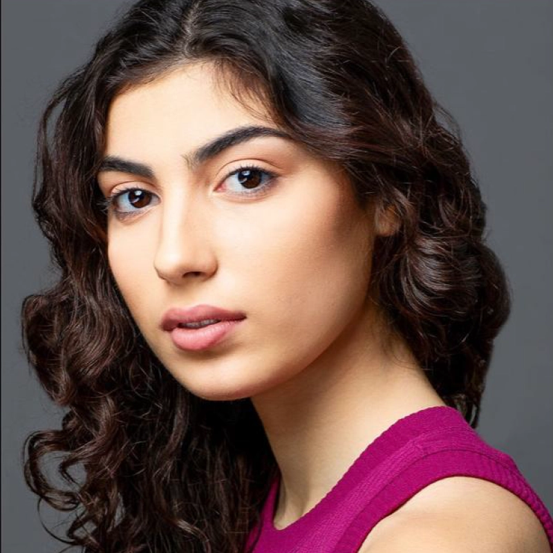 Lyla Karekinian, a young woman with long, wavy dark hair, looks slightly over her shoulder toward the camera. She has clear skin, gently arched brows, and wears a sleeveless magenta top against a plain, dark gray background. Her expression is calm and neutral.