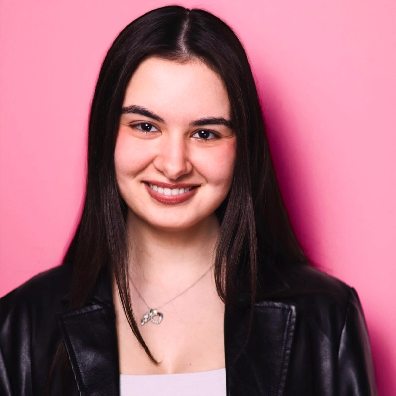 A young woman with straight, dark hair smiles at the camera. Maritina Sardis wears a black leather jacket over a white top and a silver necklace with two interlocking rings. The background behind her is solid pink.