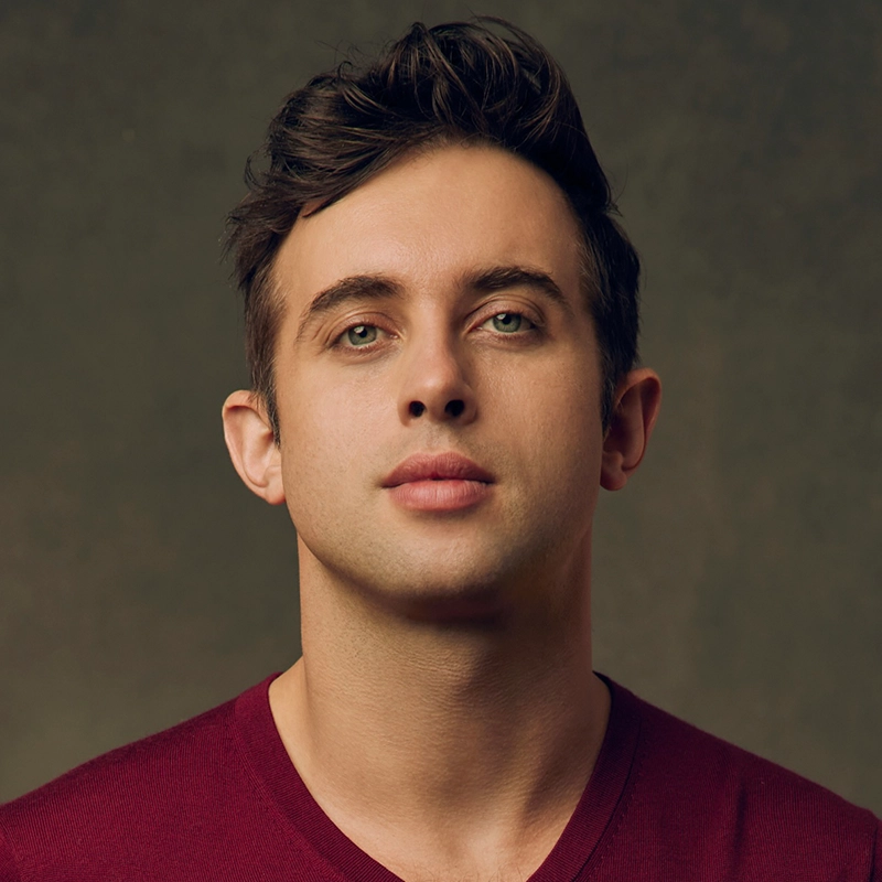 A young man with short brown hair and light green eyes faces forward against a neutral brown background. He wears a burgundy V-neck shirt and has a calm, neutral expression. The lighting is soft, highlighting his facial features.