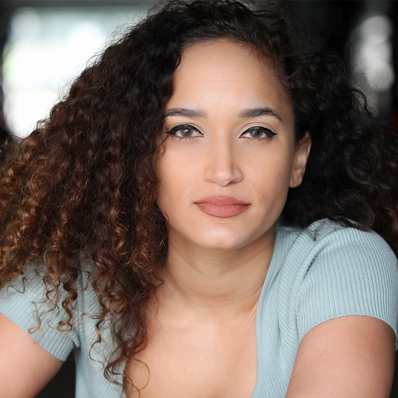 Katie Yeomans, a woman with curly brown hair and light skin, gazes confidently at the camera. She wears a light blue ribbed top and natural makeup with winged eyeliner. The blurred background keeps the focus on her expressive face.