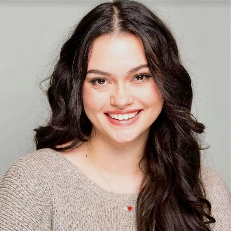 A young woman with long, wavy dark hair smiles at the camera. Ninako Donville wears a light brown knit sweater and a delicate necklace with a small red heart pendant. The plain light gray background gives the portrait a warm, friendly feel.