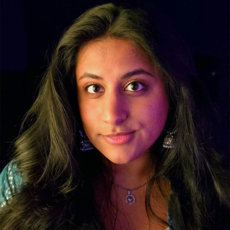 Anthara Shivkumar, a young woman with long dark hair, gazes at the camera with a slight smile. She wears large silver earrings, a pendant necklace, and a blue top. Soft, colorful lighting highlights her face against a dark background.
